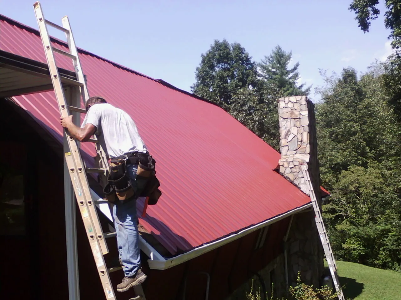 Roofer on a ladder beside a red metal roof and stone chimney — Soffit & Fascia Repair by Roofing Brothers in Allendale